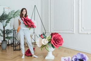 une photo horizontale d'une fleuriste fait de beaux bouquets de fleurs artificielles, se tient dans un hall spacieux, se prépare pour un événement festif, a une expression agréable. personnes, décoration et concept floral