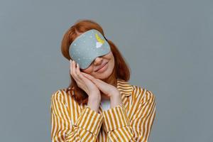 jolie femme rousse porte un bandeau sur les yeux, garde les mains jointes près du visage, voit des rêves agréables en dormant, vêtue de vêtements domestiques, isolée sur un mur gris, se sent à l'aise photo