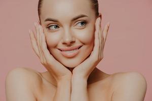 concept de lifting, de soins de la peau et de bien-être. photo de tête d'une charmante jeune femme caucasienne touche doucement les joues, a une peau hydratée éclatante, a des âmes nues, regarde tendrement de côté. portrait de beauté.