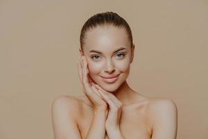 portrait de beauté naturelle. photo d'une jeune femme profite d'une peau fraîche parfaite après avoir rendu visite à un cosmétologue, sourit doucement, pose nue sur fond beige, touche le visage, porte un minimum de maquillage.