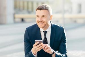 photo horizontale d'un homme séduisant avec une expression joyeuse et réfléchie, utilise un téléphone mobile moderne pour rechercher des informations sur le site Web international, connecté à Internet haut débit 4g, gagne de l'argent en ligne