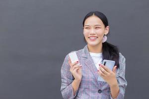 une femme asiatique professionnelle dans un blazer à rayures marron sourit joyeusement, réussie et confiante tout en tenant un smartphone et une carte de crédit sur le mur gris foncé. photo