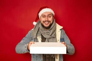 portrait d'un homme brun heureux et généreux avec une barbe en vêtements d'hiver souriant et donnant une boîte-cadeau à l'appareil photo, partageant un cadeau de vacances, concept caritatif. studio d'intérieur tourné isolé sur fond rouge photo
