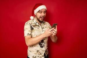 excité jeune homme touristique voyageur en vêtements d'été, bonnet de noel à l'aide d'un téléphone portable en tapant un message sms isolé sur fond rouge. passager voyageant à l'étranger en vacances concept de voyage en avion photo