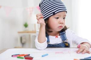 petite fille asiatique assise à table dans la chambre, fille d'âge préscolaire dessinant sur papier avec des stylos colorés le jour ensoleillé, jardin d'enfants ou photo