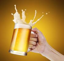 main tenant une chope de bière légère grillant avec des éclaboussures de mousse à bulles sur fond doré avec espace de copie photo