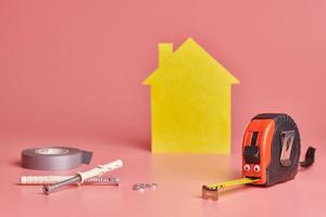 concept drôle de rénovation de maison. ruban à mesurer en métal et autres articles de réparation. réparation à domicile et concept redécoré. figure en forme de maison jaune sur fond rose. photo