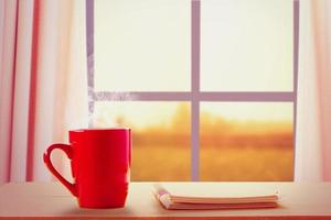 une tasse rouge de café chaud et un ordinateur portable à côté des fenêtres au lever du soleil sur fond de campagne photo