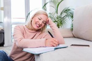 fille étudiant la veille de l'examen, écrivant des notes avec un crayon. jolie jeune fille faisant ses études à la maison assise sur le sol dans le salon avec un livre lisant un classeur de notes de cours photo