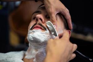 rasoir dans les mains d'un coiffeur spécialisé. coiffeur rasant un homme dans un salon de coiffure, gros plan. homme mith moustaches ayant un rasage photo