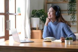 fille souriante dans les écouteurs assis à un bureau regardant à l'extérieur et utilisant un ordinateur portable pour étudier en ligne. les jeunes femmes intelligentes qui sont heureuses dans les écouteurs suivent des cours sur le Web ou s'entraînent à utiliser des ordinateurs. photo