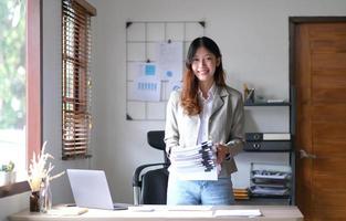 charmante femme asiatique avec un sourire debout tenant des papiers et regardant la caméra au bureau. photo