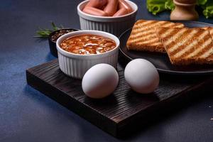 petit-déjeuner anglais traditionnel avec œufs, pain grillé, saucisses, haricots, épices et herbes sur une plaque en céramique grise photo