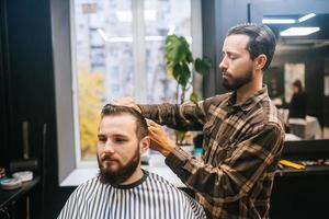 joyeux jeune homme barbu se coupe les cheveux par un coiffeur au salon de coiffure photo