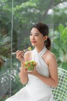 jolie femme souriante asiatique isolée sur le salon à la maison en train de manger de la salade photo