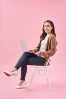 heureuse jeune femme asiatique assise sur une chaise sur fond rose, travaillant sur un ordinateur portable photo