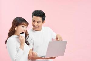 joyeux couple interracial se réjouissant du succès avec carte de crédit et ordinateur portable, regardant l'écran de l'ordinateur et criant de joie sur fond rose photo