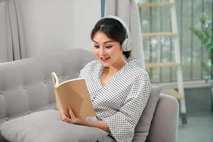 gros plan d'une femme décontractée aux cheveux bruns appréciant la musique tout en lisant un livre sur un canapé dans une maison lumineuse photo