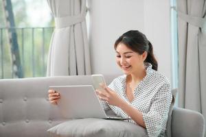 heureuse jeune femme asiatique utilisant un téléphone portable tout en étant assise sur un canapé à la maison avec un ordinateur portable photo