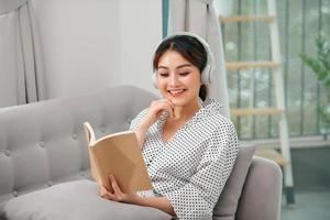 Une jeune femme asiatique séduisante se détend en se reposant à l'aide d'un téléphone intelligent pour jouer à l'application de diffusion de musique pendant la lecture d'un livre assis sur un canapé ou un canapé à la maison du jardin intérieur photo