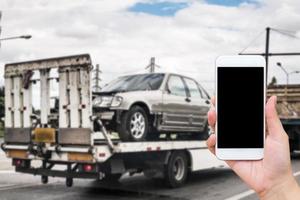 main utilisant un smartphone mobile pour un service routier d'urgence avec une voiture cassée sur une dépanneuse après un accident de la circulation photo