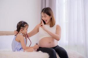 une fille asiatique utilise un stéthoscope pour sentir le ventre de sa mère pour écouter le rythme cardiaque de son bébé photo