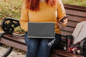 une jeune pigiste aux cheveux roux travaille dans le parc à l'aide d'un ordinateur portable. femme d'affaires moderne parlant au téléphone. travail en ligne sur internet. photo