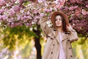 portrait de jeune belle fille à la mode au chapeau posant près d'un arbre en fleurs avec des fleurs roses par une journée ensoleillée photo