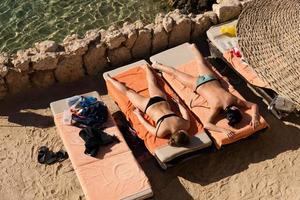 deux filles en bikini prenant un bain de soleil sur des chaises longues sur fond de mer rouge. vue d'en-haut. copines profitent du soleil, corps mince bronzé, vacances d'été, station balnéaire photo