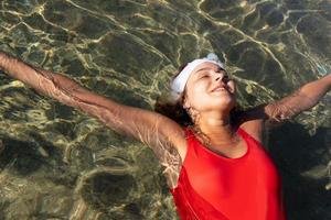 portrait en gros plan d'une belle jeune fille aux taches de rousseur, en maillot de bain rouge et chapeau de père noël, allongée dans l'eau de mer de la mer rouge. photo