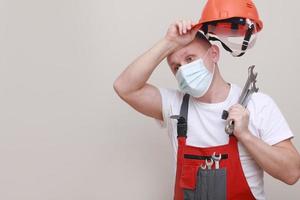 portrait d'un ingénieur fatigué ou d'un mécanicien portant un casque et un masque facial pour protéger le virus covid 19 sur fond blanc, avec une clé de maintien. photo