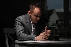 homme buvant de l'alcool dans la cuisine après le travail. jeune homme souffrant de forts maux de tête ou de migraine assis avec un verre de whisky dans la cuisine. dépendance à l'alcool photo