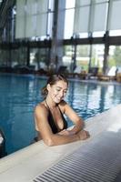 gros plan de sexy jeune femme bronzée en bikini, bord de piscine maigre, appareil photo sensuel souriant. modèle féminin attrayant en maillot de bain noir allongé dans l'eau de la piscine à l'hôtel, concept de tourisme.
