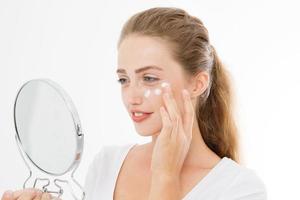 jeune femme blonde caucasienne avec miroir et crème hydratante pour les soins de la peau du visage sur le visage isolé sur fond blanc. concept anti-âge et spa. maquillage et beauté de la nature. copie espace photo