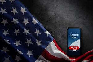 jour des élections dans le concept des états-unis. spectacle de vote en ligne sur l'écran du téléphone mobile. smartphone allongé sur fond de ciment et drapeau américain à proximité. vue de dessus photo
