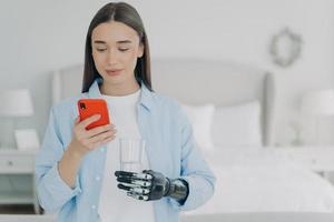 une jeune fille handicapée tient un smartphone et un verre d'eau dans les mains à l'aide d'une prothèse bionique de haute technologie photo