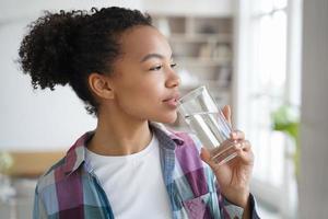 une jeune fille métisse boit de l'eau pure du verre à la maison. mode de vie sain, routine du matin photo