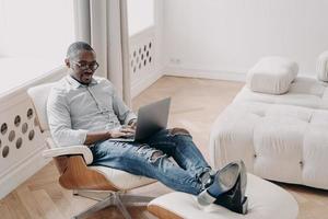 homme afro-américain travaillant en ligne sur un ordinateur portable, assis dans un fauteuil au bureau à domicile. travail à distance photo