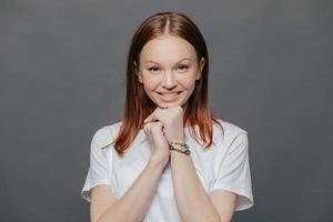 photo horizontale d'une belle femme ravie porte un t-shirt blanc décontracté, garde les mains sous le menton, sourit doucement, est de bonne humeur, pose sur fond de studio gris foncé. Émotions positives