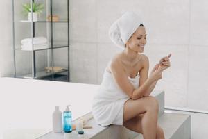 une femme souriante enveloppée dans une serviette contient un échantillon de cosmétiques assis dans la salle de bain. soins de la peau, spa photo