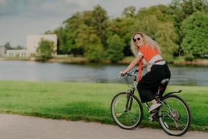 photo d'une femme gaie habillée avec désinvolture, fait du vélo, regarde de côté, a une expression heureuse, porte des nuances, pose près de la rivière, de la pelouse verte et des arbres, quelques bâtiments en arrière-plan. les gens et le repos