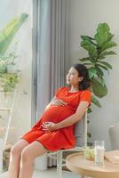 fière femme enceinte regardant son ventre assise sur un canapé dans le salon à la maison avec une lumière chaude entrant par la fenêtre photo