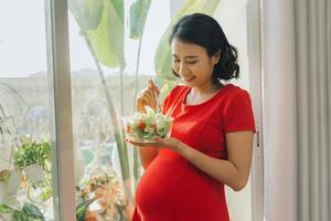 jeune femme enceinte mangeant une salade de légumes près de la fenêtre à la maison photo