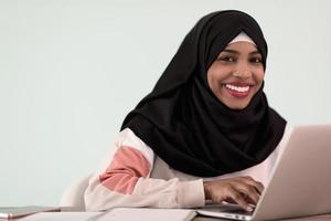 une femme musulmane afro portant un hijab est assise en souriant dans son bureau à domicile et utilise un ordinateur portable photo