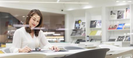 portrait d'asain jolie femme gaie confortable assise et lisant un livre dans la bibliothèque et s'appuyant sur un mur de verre avec plaisir, concentration et concentration sur le contenu photo