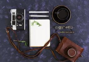 table de bureau à plat, vue de dessus. espace de travail de bureau avec appareil photo rétro, agenda, stylo, lunettes, étui, tasse de café, romarin sur fond sombre.