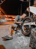 un vieil homme en short court s'efforce de changer le pneu arrière de la moto la nuit dans son atelier. photo prise la nuit à l'extérieur dans le village de luhu, gorontalo le 21 août 2022
