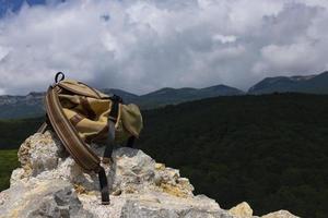 un sac à dos brun-vert repose sur une grosse pierre grise sur un rocher sur fond de montagnes verdoyantes. photo
