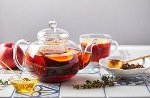 thé aux fruits avec pommes et thym dans une théière en verre et une tasse sur une table en carreaux de couleur