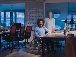 homme d'affaires en fauteuil roulant dans un espace de bureau de coworking moderne travaillant tard dans la nuit au bureau. collègues en arrière-plan. concept d'invalidité et de handicap. photo